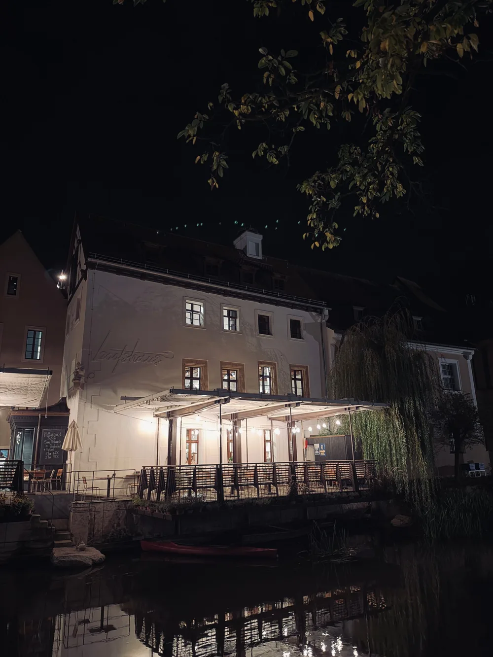 A lit terrace by the water at night, in front of a white building with a canoe moored below.