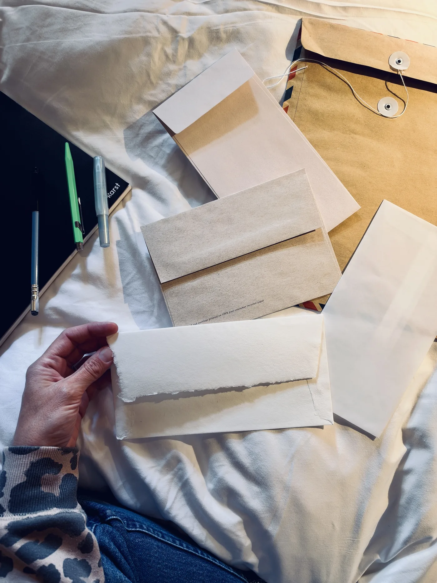Candidate envelopes laid out on a white sheet, one with a deckle edge, one recycled brown, next to pens and a notebook.