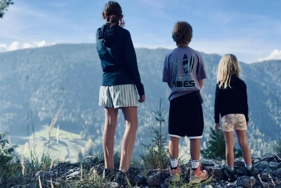 Three children seen from behind, looking out over a forested valley.