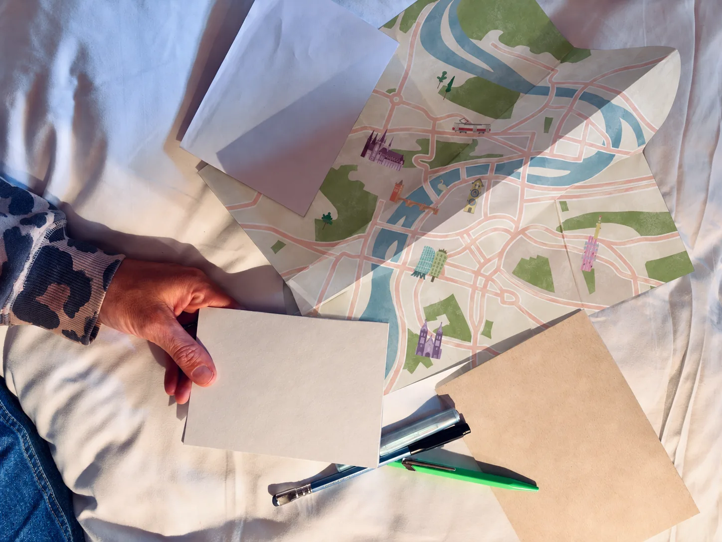 A hand holding a blank zine cover next to a folded city map and a blank postcard, on a bed.