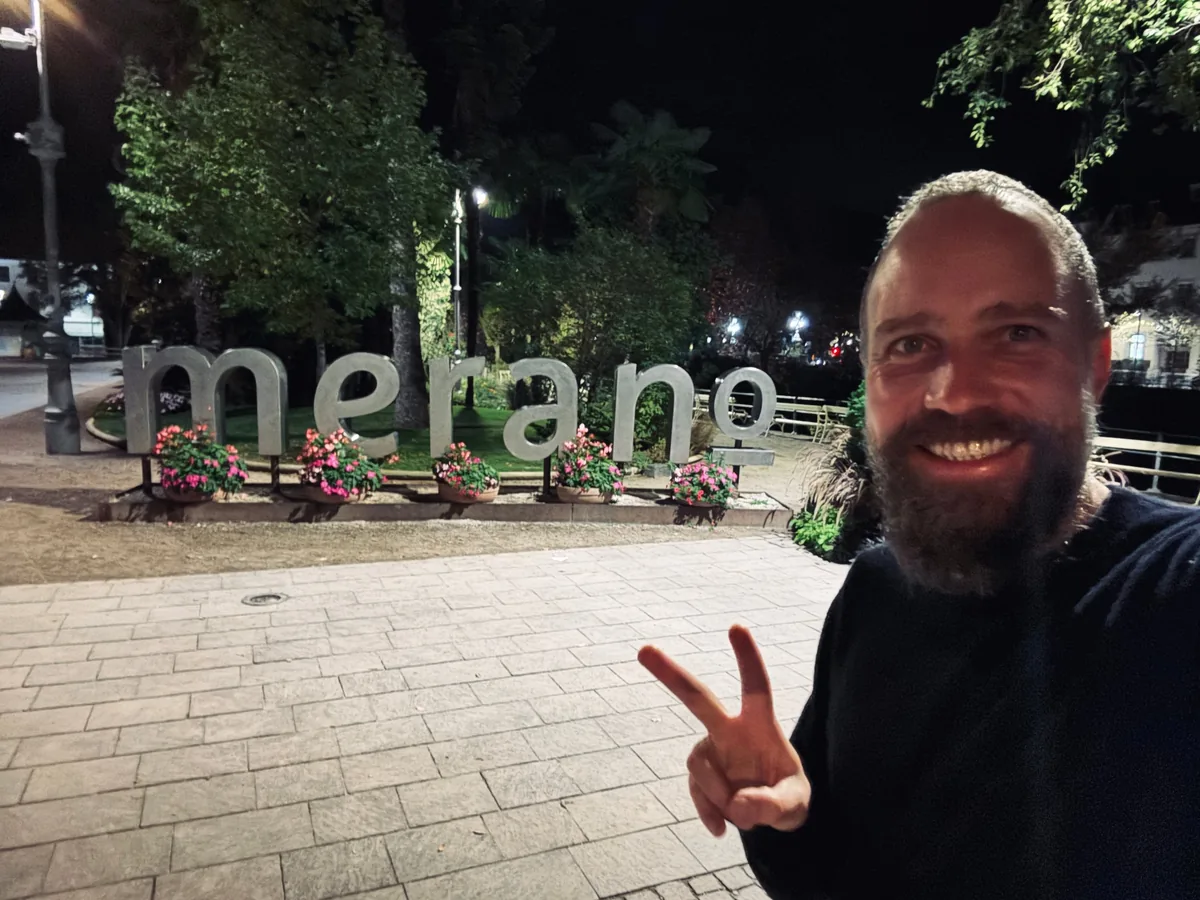 Jakob at night in front of the illuminated Merano town sign, making a peace sign.