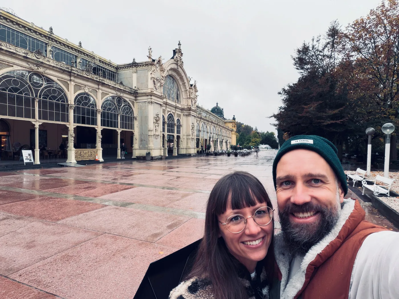Sina and Jakob taking a selfie in the rain in front of a long white colonnade in Czechia.